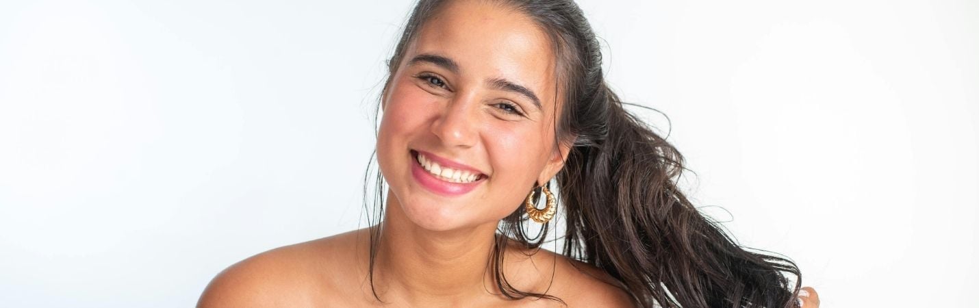 Mulher jovem com cabelo castanho-escuro preso num rabo de cavalo, sorridente, usando brincos dourados grandes, sobre fundo branco.