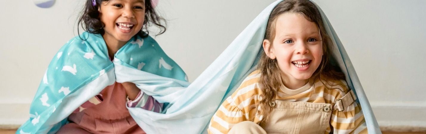 Duas crianças sorridentes sentadas no chão, cobertas com mantas azuis com padrões de coelhos, a brincar num ambiente interior decorado com ovos coloridos colados na parede.