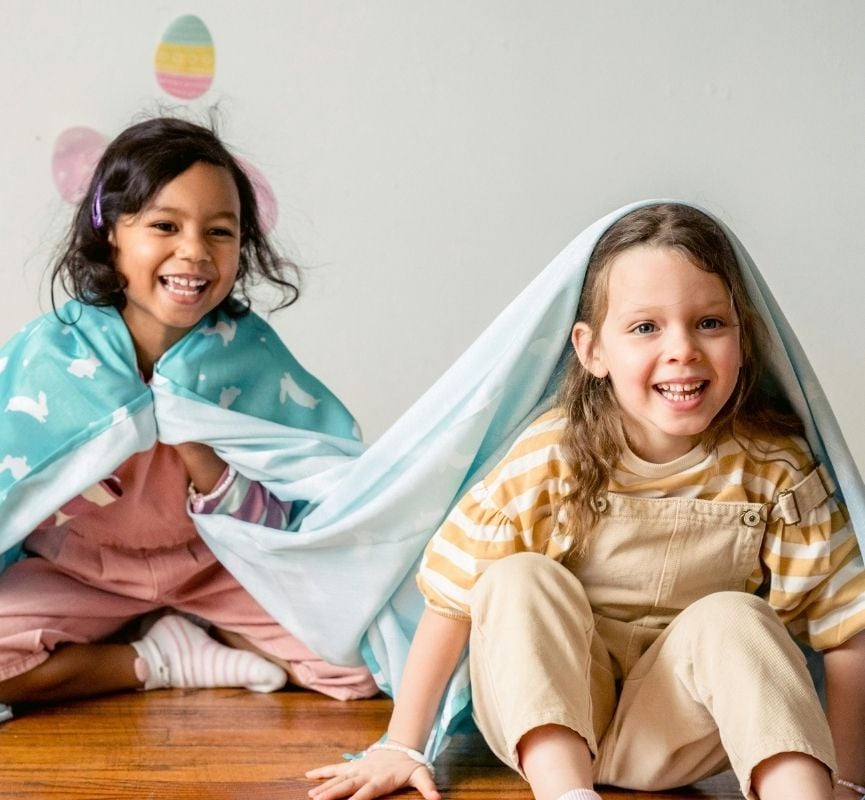 Duas crianças sorridentes sentadas no chão, cobertas com mantas azuis com padrões de coelhos, a brincar num ambiente interior decorado com ovos coloridos colados na parede.