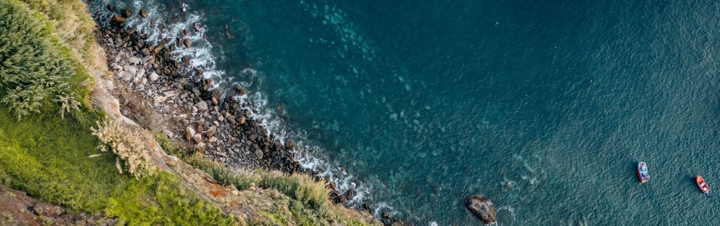 Vista aérea de uma costa com mar azul e vegetação verde, com destaque para um banner promocional a anunciar a campanha do Dia do Ambiente e dos Oceanos da Pegada Verde: “De 5 a 8 de junho – 20% em tudo”. Campanha ecológica com foco na sustentabilidade.