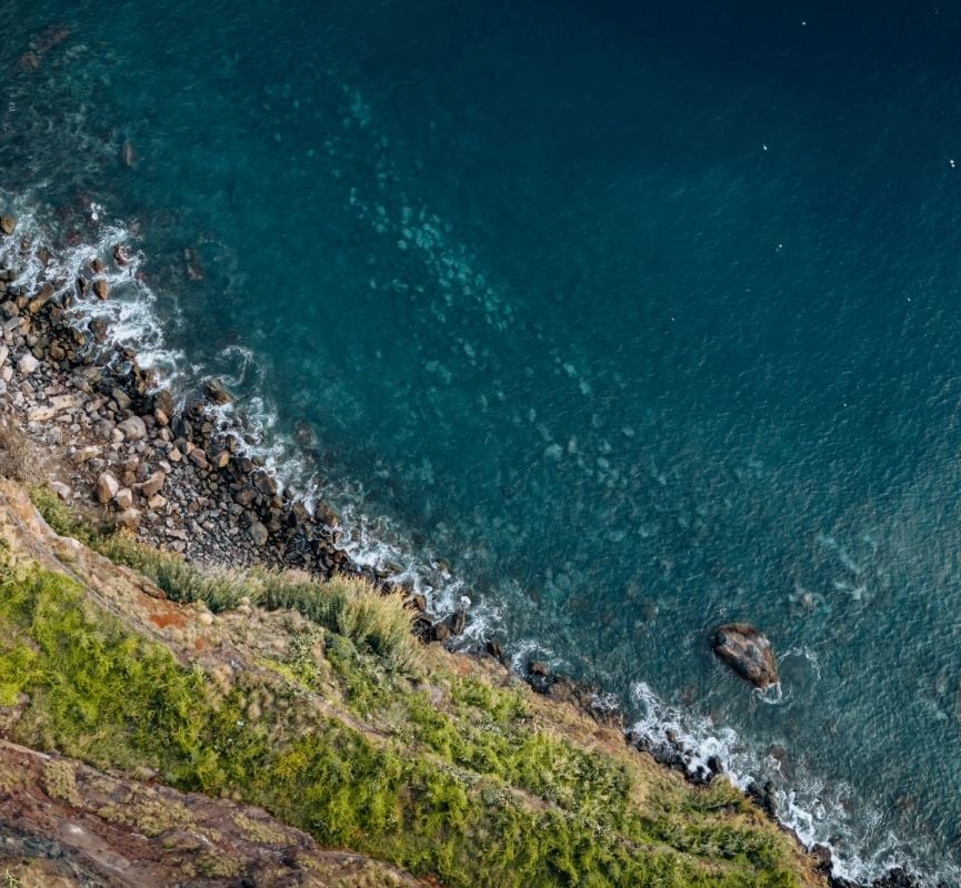 Vista aérea de uma costa com mar azul e vegetação verde, com destaque para um banner promocional a anunciar a campanha do Dia do Ambiente e dos Oceanos da Pegada Verde: “De 5 a 8 de junho – 20% em tudo”. Campanha ecológica com foco na sustentabilidade.
