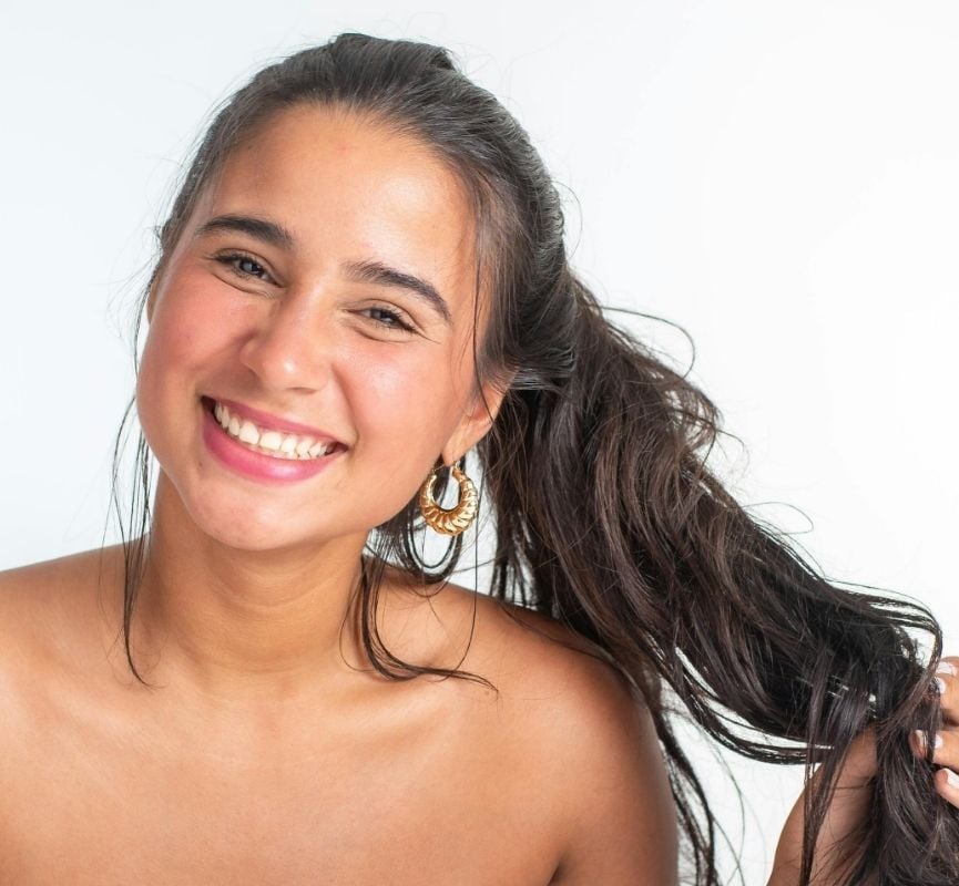 Mulher jovem com cabelo castanho-escuro preso num rabo de cavalo, sorridente, usando brincos dourados grandes, sobre fundo branco.